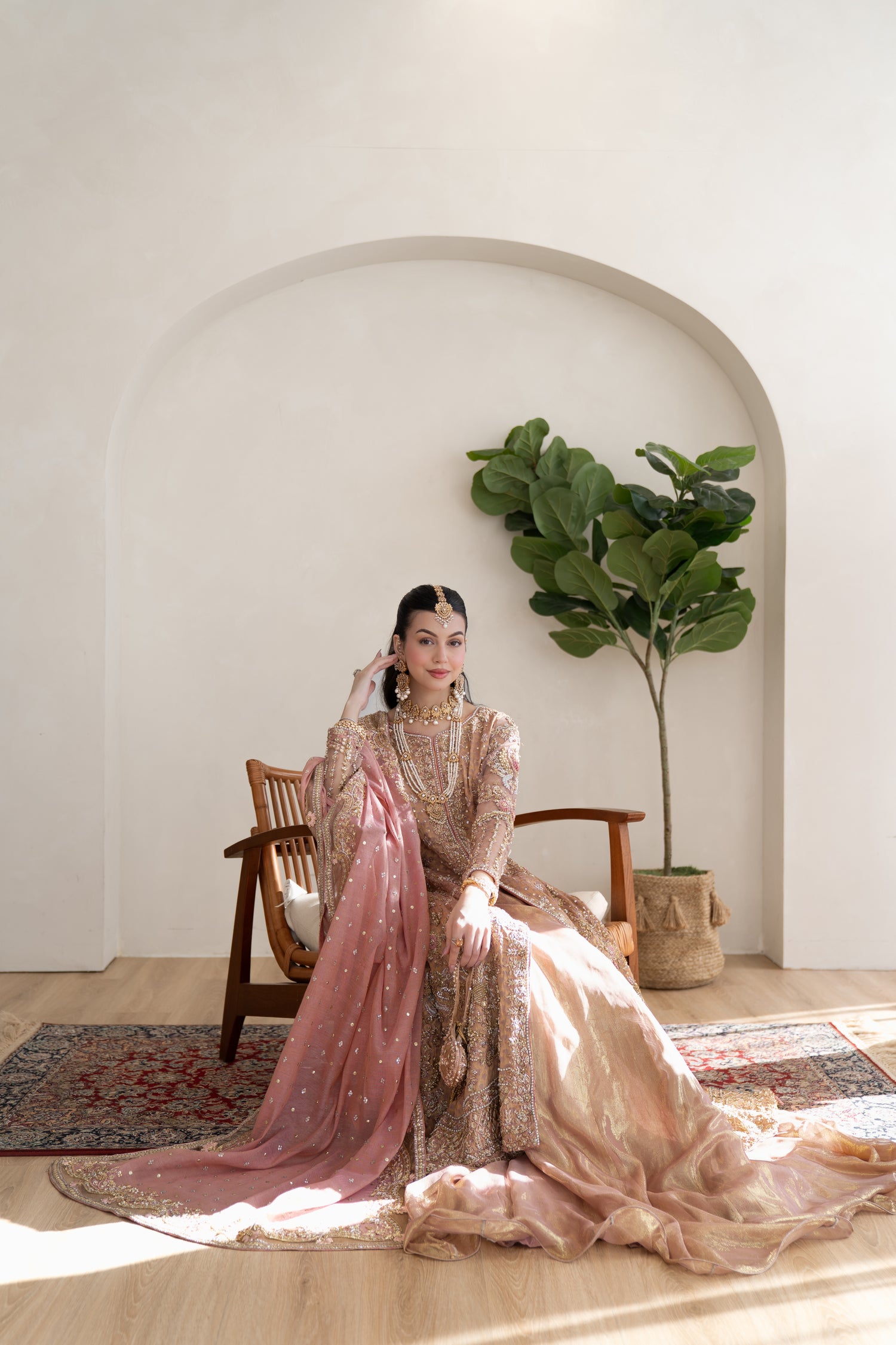 Woman in a traditional outfit sitting in a room with a plant and wooden chair.